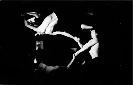 Kilku wysportowanych aktorów bez koszulek trzyma się za ręce i wykonuje akrobatyczne pozy. Aktor po prawej stronie ma skupiony wyraz twarzy.