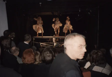 Na małej scenie tańczą trzy osoby aktorskie. Trudno określich ich płeć. Ubrane są białe krótkie sukienki. Na głowach mają blond peruki. Pod sceną siedzi publiczność zwrócona plecami w stronę widza. Pośród publiczności wyróżnia się jeden meżczyzna w okularach patrzący w obiektyw.