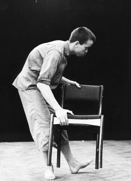 Przygarbiony aktor trzyma w rękach lekko uniesione krzesło. Ubrany jest w jasne spodnie i jasną koszulę. Jest boso.