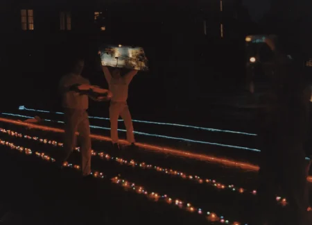 Niewyraźne zdjęcie. Noc. Przestrzeń miejska. Noc. Przestrzeń miejska. Na piasku leżą trasy wyznaczone przez czerwone i niebieskie lampki. Pośród lampek stoją aktorzy ubrani na biało. Jeden aktor trzyma nad głową obraz przedstawiający niebo i drzewa.