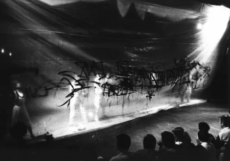 Grupa ledwo widocznych aktorów i aktorek stoi za przeźroczystą płachtą. Na płachcie widnieją nieczytelne napisy namalowane farbą. Na pierwszym planie sidzi zwrócona plecami do widza publiczność oglądająca aktorów.