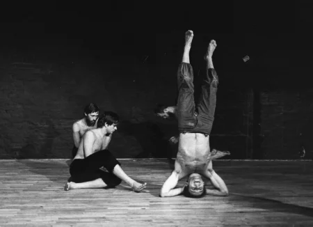 W centrum, na pierwszym planie aktor stoi na głowie. Ubrany jest w ciemne spodenki, ma nagi tors, jest boso. Głowę zwróconą ma w stronę widza. Po jego lewej stronie siedzi dwóch aktorów ubranych podobnie jak on. Patrzą na niego. Za aktorem stojącym na głowie słabo widoczny aktor wykonuje ruch akrobatyczny.