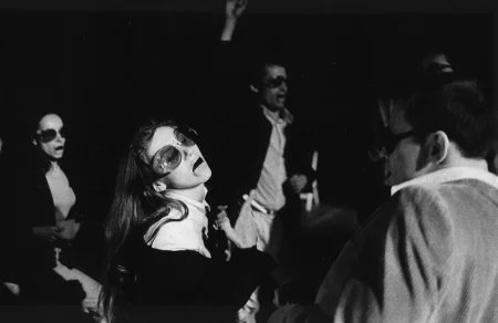 Przestrzeń miejska. Między budynkami stoi tłum ludzi zwróconych twarzą w stronę widza. Niektórzy trzymają w rękach białe flagi.