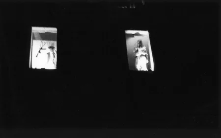 Niewyraźne zdjęcie. Dwa okna zaciemnionego budynku. W każdym oknie stoi jeden aktor zwrócony twarzą w stronę widza.