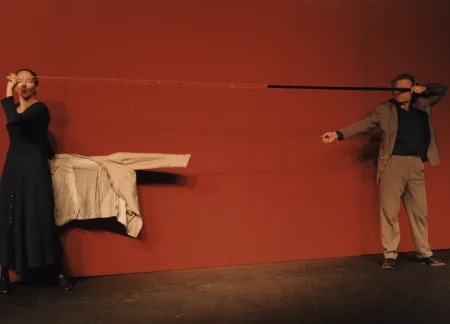 Na tle czerwonej ściany stoi aktorka i aktor. Aktorka stoi z lewej strony, aktor z prawej. Trzymają w rękach długą taśmę, z lewej strony przeźroczystą, z prawej czarną. Aktorka ubrana jest w granatową sukienkę. Za nią na lince wisi biała marynarka. Aktor ubrany jest w szary garnitur i granatową koszulę. Jego oczy zasłania czarny fragment taśmy.