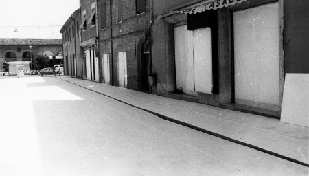 Ptrzestrzeń miejska. Ulica między budynkami jest pusta. Drzwi i okna budynków zasłonięte są białymi płachtami.