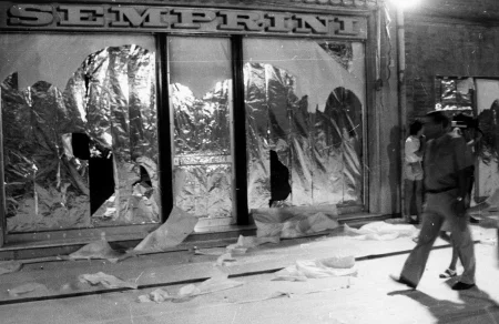 Przestrzeń miejska. Ulica wyglądająca jak tuż po zamieszkach. Pod zasłoniętymi witrynami sklepowymi leżą białe flagi. Nad drzwiami budynki widnieje napis "SEMPRINI". Z prawej strony idzie przechodzień.