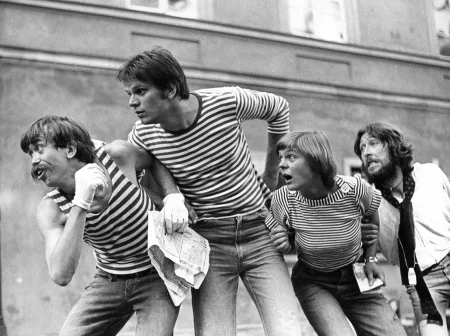 Trzech aktorów i jedna aktorka. Stoją w szeregu, trzymają się pod ręce i patrzą ze zdziwieniem w bok. Dwóch aktorów i jedna aktorka ubrani są w jeansy i podkoszulki w czarno-białe paski. Aktor z prawej strony ubrany jest w jeansy i białą koszulę. Za nimi stoi blok mieszkalny.