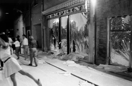 Przestrzeń miejska. Ulica wyglądająca jak tuż po zamieszkach. Pod zasłoniętymi witrynami sklepowymi leżą białe flagi. Ulicą idzie tłum ludzi.