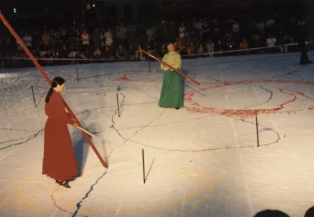 Scena ustawiona w przestrzeni miejskiej. Scena jest biała, pełna kolorowych linii. Na scenie stoi aktorka i aktor. Aktorka ubrana jest w czerwoną suknię. Aktor ubrany jest w zieloną spódnicę i jasnozieloną koszulę. Oboje trzymają w rękach długie czerwone kije, z których wysypuje się proszek. W tle widać publiczność.