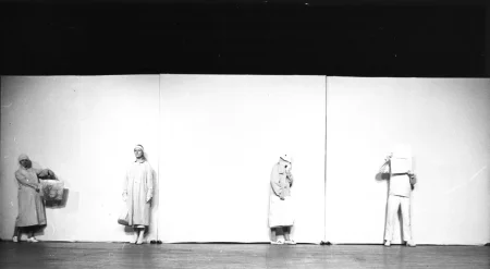 Czterosobowa grupa aktorów i aktorek stoi pod białą ścianą. Są ubrani na biało, przez co wydają się zlewać ze ścianą. Aktorka z lewej strony trzyma w rękach torbę i podnosi ją. Aktorka obok niej stoi na baczność i patrzy w stronę widza. Obok niej stoi aktorka zasłaniająca twarz ubraniami. Po prawej stronie stoi aktor zasłaniający twarz gazetą.