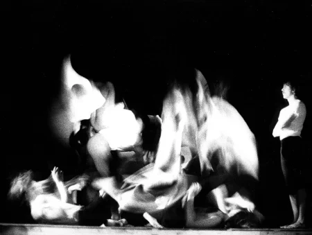 Niewyraźne, rozmazane zdjęcie. Grupa aktorów skacze i leży na pustej scenie. Po prawej stronie stoi aktor ubrany w biały podkoszulek i czarne spodenki. Jest bosy. Patrzy na grupę aktorów nie wyrażając emocji.