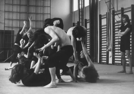 Grupa aktorów i aktorek w sali gimnastycznej. Kłębią się na podłodze. Są ubrani w czarne spodenki i czarne podkoszulki. Niektórzy mają nagie torsy. Są boso. Po prawej stronie stoi aktor przyglądający się grupie. Ma czarne spodenki i czarny podkoszulek. Jest boso.