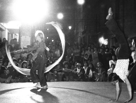 Scena wśród budynków. Aktorka po lewej stronie, zwrócona profilem do widza, macha białą wstęgą. Aktorka po prawej stronie stoi na rękach. Na drugim tle, na placu miejskim widać widzów oglądających aktorki.