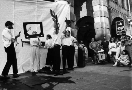 Przestrzeń miejska. Pod białą płachtą zawieszoną na kolumny budynku stoi sześcioosobowa grupa aktorów i aktorek. Czterech aktorów ubranych jest w czarne spodnie i białe koszule. Jedna aktorka ubrana jest w jasne spodnie i białą koszulę. Druga aktorka ubrana jest w czarną spódnicę i białą koszulę. Wszyscy trzymają w rękach dwuwymiarowe czarne rekwizyty wyglądające na wycięte z papieru, takie jak telewizor, piła do cięcia drewna i domek. Po prawej stronie stoją widzowie oglądający aktorów. Za ich plecami stoi budynek.