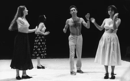 Na scenie stoją trzy aktorki i jeden aktor. Aktorki ubrane są w spódnice i staniki. Aktor jest ubrany w jasne spodnie i ma nagi tors. Wszyscy gestykulują dłońmi.