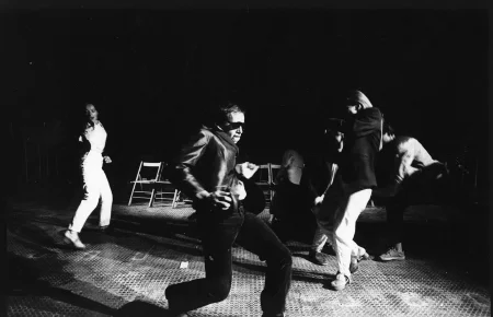 Przestrzeń miejska. Ulica wyglądająca jak tuż po zamieszkach. Pod zasłoniętymi witrynami sklepowymi leżą białe flagi. Ulicą idzie tłum ludzi.