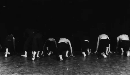 Grupa aktorów i aktorek zwróconych plecami w stronę widza. Wyglądają, jakby przygotowywali się do biegu. Aktorki ubrane są na czarno. Aktorzy ubrani są w czarne spodenki i mają nagie torsy. Wszyscy są boso.