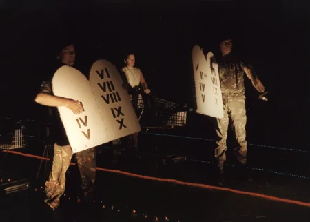 Noc. Na piasku leżą trasy wyznaczone przez czerwone i niebieskie lampki. Pośród lampek stoją aktorzy i wózki sklepowe. Dwóch aktorów w ubraniach wojskowych trzyma tablice z rzymskimi cyframi kończącymi się na "X". Tablice przypominają Dekalog. Aktorzy odrywają w tablic kolejne cyfry i rzucają je na ziemię.