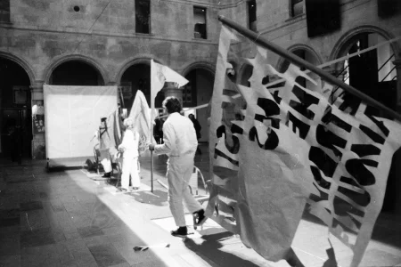 Plac wewnątrz budynku. Na podłodze leży rozwinięty wybieg. Wzdłuż wybiegu stoi grupa aktorów i aktorek odwróconych do widza plecami. Ubrani są na biało i trzymają w rękach flagi. Na pierwszym planie wisi flaga z nieczytelnymi napisami. W tle widać kolumny budynku.