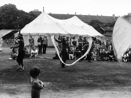 Plener, ruiny jakiegoś budynku. W centrum na czarno ubrana aktorka macha białą wstęgą. W tle siedzą i stoją widzowie, którzy oglądają aktorów. Na pierwszym planie widać dziecko odwrócone plecami do widza. Dziecko ogląda działania aktorskie. Na dalszym planie stoi namiot cyrkowy.