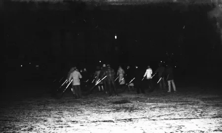 Niewyraźne zdjęcie. Przestrzeń miejska. Na trawniku stoi grupa aktorów i aktorek zwróconych do widza plecami. Trzymają w rękach miotły. W oddali widać budynek.