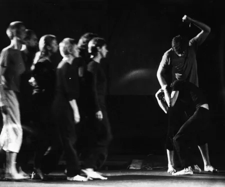 Po lewej stronie grupa aktorów i aktorek zwrócone do nas profilem w szeregu idzie w prawą stronę. Są ubrani w czarne spodenki i czarne podkoszulki. Są boso. Po prawej stronie stoi dwoje aktorów. Jeden jest niższy, drugi wyższy. Wyższy aktor bije niższego. Niższy jest skulony.