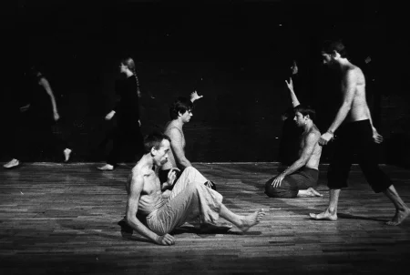 Po lewej stronie idą w szeregu zwrócone profilem do widza trzy aktorki. Są ubrane na czarno, są boso. W centrum, na pierwszym planie siedzi aktor. Ubrany jest w jasne spodenki, ma nagi tors. Jest boso. Jest zgarbiony i patrzy w podłogę. Za nim klęczy dwóch aktorów Ubrani są w ciemne spodenki, mają nagie torsy. Klęczą naprzeciwko siebie, każdy z nich ma jedną rękę wyciągniętą do boku. Z prawej strony idzie, w tym samym kierunku co aktorki, idzie aktor. Ubrany jest w czarne spodenki, ma nagi tors. Jest boso.