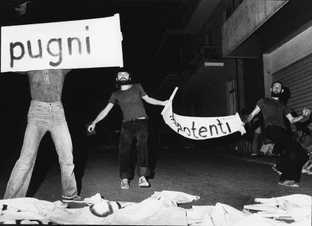 Trzech aktorów stoi na ulicy przodem do widza. Aktor z lewej strony trzyma zasłaniający mu głowę transparent z napisem "pugni". Ubrany jest w jeansowe spodnie. Po prawej stronie stoi dwóch aktorów trzymających transparent z napisem, który trudno rozczytać. Ubrani są w jeansowe spodnie i ciemne podkoszulki. W tle widać budynki mieszkalne z balkonami.