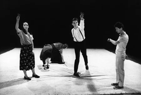 Na białej płachcie stoi grupa aktorów i aktorek. Aktor i aktorka patrzą w stronę widza i unoszą jedną dłoń. Dwie aktorki z tyłu są schylone i dotykają dłońmi białej płachty. Z prawej strony stoi zwrócony profilem w stronę widza aktor patrzący na zegarek.