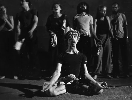 W centrum siedzi aktor w siadzie skrzyżnym. Wygląda, jakby medytował. Trzyma zgięte ręce po bokach, ma otwarte usta. Widać tylko jego białka oczu. Na głowie ma arafatkę. Za nim w szregu stoi grupa aktorów i aktorek. Wyglądają, jakby śpiewali.