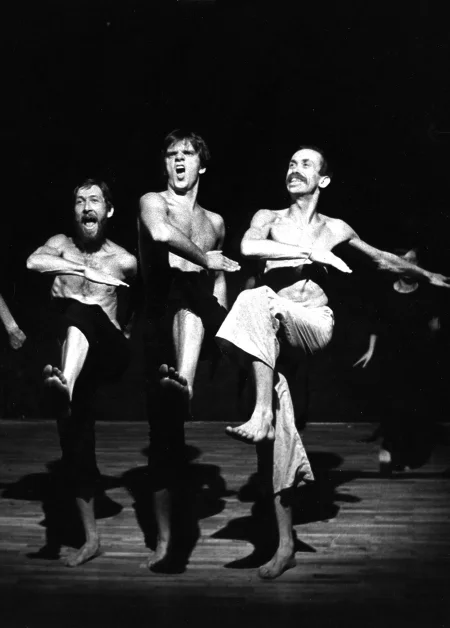 Trzech aktorów w szeregu, zwróconych przodem do widza wygląda, jakby energicznie maszerowało. Aktorzy szeroko się uśmiechają. Ubrani są w spodnie, mają nagie torsy. Są boso.