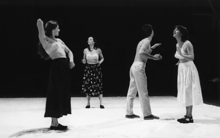 Na scenie stoją trzy aktorki i jeden aktor. Aktorki ubrane są w spódnice i staniki. Aktor jest ubrany w jasne spodnie i ma nagi tors. Wszyscy gestykulują dłońmi.