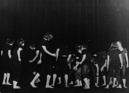 Liczna grupa aktorów w czarnych podkoszulkach i czarnych spodenkach bez butów. Są odwróceni do nas tyłem, wyglądają jakby tańczyli. Z prawej strony stoją dwie aktorki zwrócone do siebie twarzami. Jedna z nich jest zwrócona do nas tyłem, druga przodem.