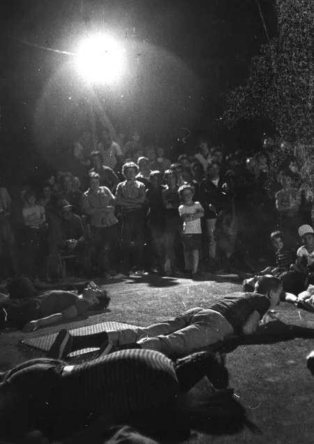 Plener wieczorem. Grupa aktorów i aktorek leży na ziemi - niektórzy brzuchem do dołu, inni plecami. Aktorom i aktorkom przygląda się grupa widzów. Wśród widzów stoi lampa oświetlająca aktorów i aktorki.