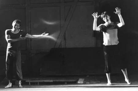 Dwóch aktorów stoi w industrialnej przestrzeni. Aktor po lewej stronie ubrany jest w czarne spodnie i czarne odkoszulek. Jest bosy. Uśmiecha się w stronę widza i wskazuje rękami na aktora stojącego po prawej stronie. Aktor po prawej stronie ubrany jest w czarne spodenki. Jego koszulka jest czarno-biała, choć może to być kwestia oświetlenia. Jest bosy. Uśmiecha się w stronę widza. Ma podniesione ręce, pokazuje wewnętrzną część obu dłoni.