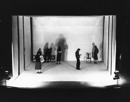 Minimalistyczna scena pudełkowa pomalowana na biało. Po dwóch stronach tylnej ściany stoi ławka i krzesło. Scena oświetalna jest dwoma reflektorami. Na scenie stoi sześcioosobowa grupa aktorów i aktorek. Nie zwacają na siebie uwagi. Rzucają cienie na tylną ścianę.