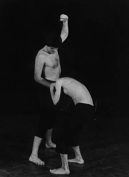 Dwóch aktorów stoi na pustej scenie. Są boso, mają czarne spodenki i nie mają koszulek. Aktor po prawej ma wyciągniętą do góry jedną rękę. Drugą ręką trzyma drugiego aktora za włosy. Wygląda, jakby go bił. Drugi aktor jest skulony.