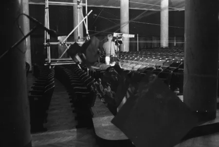 Pusta widownia teatralna widziana z boku. Na ścianach wisi pełno linek. Wśród krzeseł stoi zwrócony plecami w stronę widza mężczyzna.