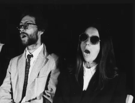 Ptrzestrzeń miejska. W małej białej kabinie stoi aktorka. Jest ubrana na biało i ma mikrofon przy twarzy. Obok kabiny przechodzi kobieta z wózkiem. Na ulicy stoi grupa ludzi.