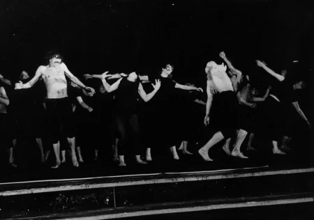 Grupa aktorów i aktorek stoi na scenie. Ich ciała są powyginane. Niektórzy mają na sobie czarne spodenki i czarne koszulki, inni nie mają na sobie koszulek. Wyglądają, jakby cierpieli.