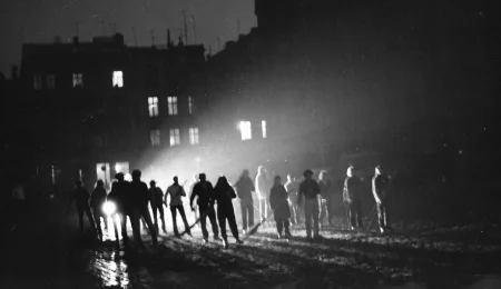 Niewyraźne zdjęcie. Przestrzeń miejska. Między blokami stoi grupa aktorów i aktorek. W rękach trzymają coś, co przypomina miotły. Za ich plecami leżą reflektory, które oświetlają przestrzeń.