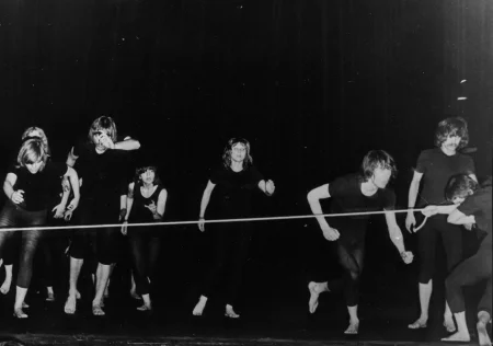 Grupa aktorów i aktorek ubrana w czarne koszulki i czarne spodenki, bez butów. Biegną przed siebie, zwróceni do nas przodem. Na ich drodze stoi napięta lina, którą trzyma aktor stojący po prawej stronie.