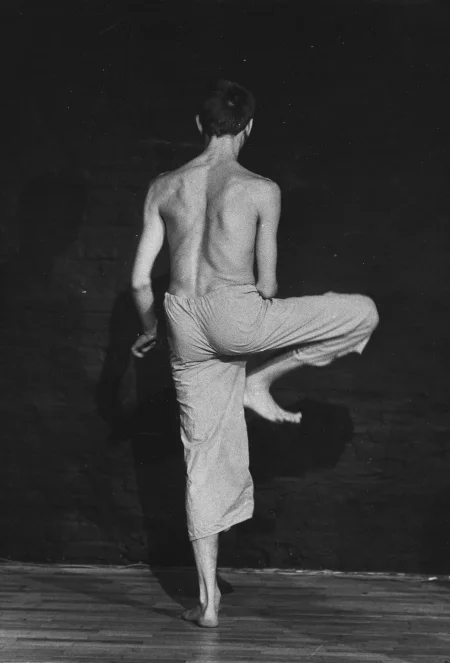 Aktor stoi zwrócony plecami do widza. Ubrany jest w jasne spodenki, ma nagi tors. Jest boso. Stoi na jednej nodze. Jego druga noga jest zgięta w kolanie.