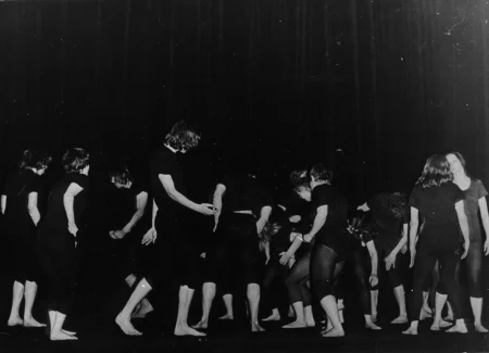 Liczna grupa aktorów w czarnych podkoszulkach i czarnych spodenkach bez butów. Są odwróceni do nas tyłem, wyglądają jakby tańczyli. Z prawej strony stoją dwie aktorki zwrócone do siebie twarzami. Jedna z nich jest zwrócona do nas tyłem, druga przodem.