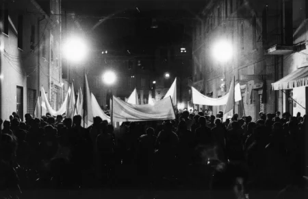 Przestrzeń miejska. Między budynkami stoi tłum ludzi zwróconych plecami w stronę widza. Niektórzy trzymają w rękach białe flagi.