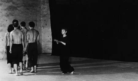 Po lewej stronie sceny na tle czarnej płachty stoi grupa aktorów odwróconych do widza plecami. Ubrani są w ciemne spodenki, mają nagie torsy. Są boso. W centrum stoi aktorka ubrana w czarne spodnie i czarną koszulkę. Jest boso. Ma krótkie włosy. Gestykuluje dłońmi.