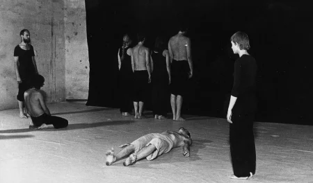 Grupa aktorów i aktorek na tle czarnej płachty. Po obu stronach naprzeciwko siebie stoją aktor i aktorka. Oboje są ubrani na czarno. Obok aktora klęczy aktor. Ubrany jest w czarne spodenki, jego tors jest nagi. Pod płachtą, plecami w stronę widza stoją aktorzy i aktorki. Aktorzy ubrani są w czarne spodenki, mają nagie torsy. Aktorki ubrane są w czarne spodnie i czarne koszulki na ramiączka. W centrum na plecach leży aktor. Ubrany jest w jasne spodenki, jego tors jest nagi.