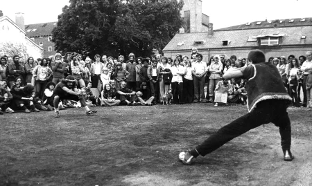 Plener. Aktor zwrócony do widza plecami ma ugiętą jedną nogę. Ubrany jest w czarne spodnie i ciemną kamizelkę. Patrzy w stronę pokaźnej grupy widzów. W tle widać drzewa i budynki.