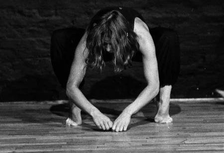 Aktorka, mocno pochylona w stronę podłogi, kuca stojąc na palcach. Dłońmi lekko dotyka podłogi. Ubrana jest w czarne spodnie i czarną koszulkę na ramiączka. Jest boso.
